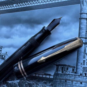 May include: A black fountain pen with gold accents, the nib exposed, and the cap partially open. The pen is set against a backdrop of a blue-toned image of a bridge.