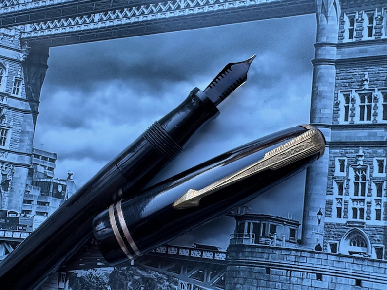 May include: A black fountain pen with gold accents, the nib exposed, and the cap partially open. The pen is set against a backdrop of a blue-toned image of a bridge.