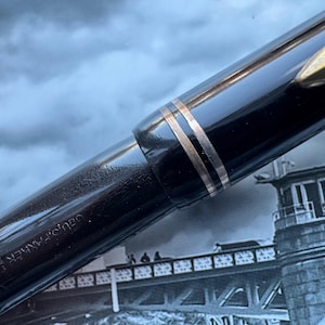 May include: Close-up of a black fountain pen with gold-colored accents. The pen has a shiny, reflective surface and a gold-colored clip. The pen's body is engraved with the brand name. The background is a blurred image of a bridge and buildings.