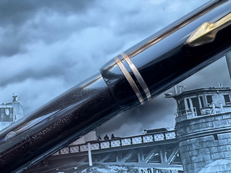 May include: Close-up of a black fountain pen with gold-colored accents. The pen has a shiny, reflective surface and a gold-colored clip. The pen's body is engraved with the brand name. The background is a blurred image of a bridge and buildings.
