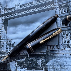 May include: A black fountain pen with gold accents, including a gold nib and clip, and a matching cap. The pen is set against a black and white backdrop of a bridge.