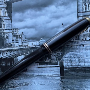 May include: A black fountain pen with gold-colored accents. The pen is set against a backdrop of a black and white image of a bridge and buildings, creating a classic and elegant aesthetic. The pen's design suggests a focus on writing instruments.
