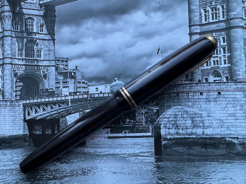 May include: A black fountain pen with gold-colored accents. The pen is set against a backdrop of a black and white image of a bridge and buildings, creating a classic and elegant aesthetic. The pen's design suggests a focus on writing instruments.