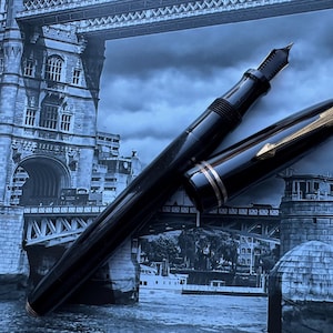 May include: A black fountain pen with gold accents, including a gold nib and a gold band near the cap. The pen is set against a backdrop of a black and white image of a bridge.
