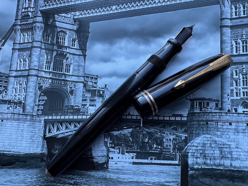 May include: A black fountain pen with gold accents, including a gold nib and a gold band near the cap. The pen is set against a backdrop of a black and white image of a bridge.