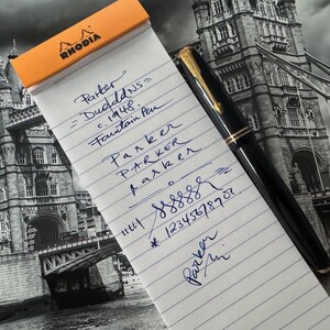 May include: A black fountain pen with gold accents rests on a notepad with the word "RHODIA" at the top. The notepad has handwritten text in blue ink, including the words "Parker" and "Fountain Pen". The background features a black and white image of a bridge.