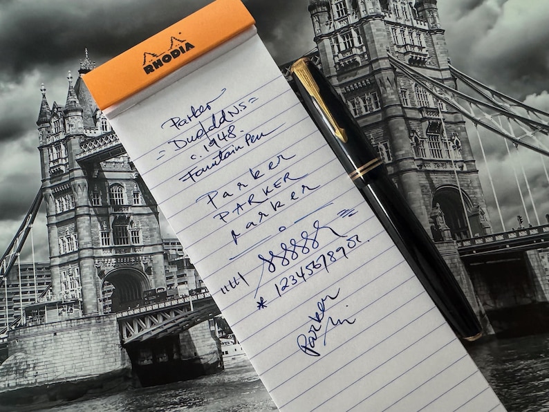 May include: A black fountain pen with gold accents rests on a notepad with the word "RHODIA" at the top. The notepad has handwritten text in blue ink, including the words "Parker" and "Fountain Pen". The background features a black and white image of a bridge.