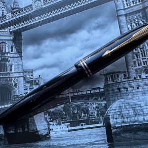 May include: A black fountain pen with gold-colored accents, including the clip and bands, is displayed against a backdrop of the Tower Bridge in London. The pen has a sleek, polished appearance.