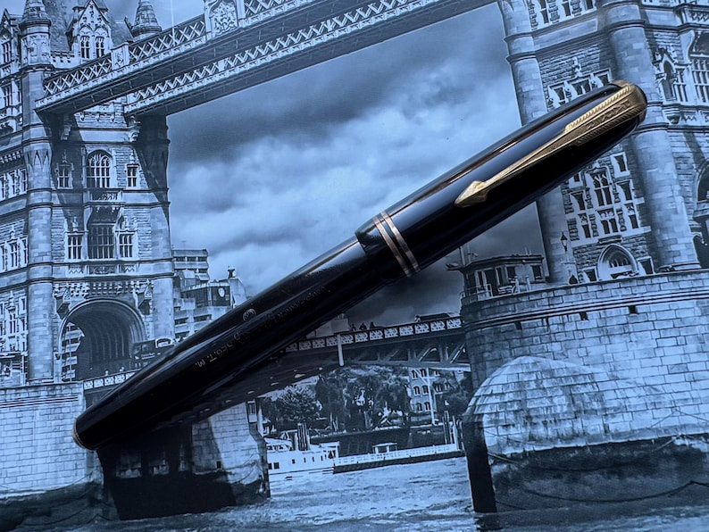 May include: A black fountain pen with gold-colored accents, including the clip and bands, is displayed against a backdrop of the Tower Bridge in London. The pen has a sleek, polished appearance.