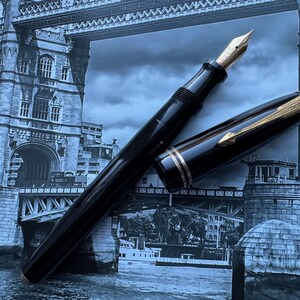 May include: A black fountain pen with a gold nib and accents, lying open on a background of the Tower Bridge in London. The pen's cap is off, revealing the nib. The pen has a sleek, classic design.