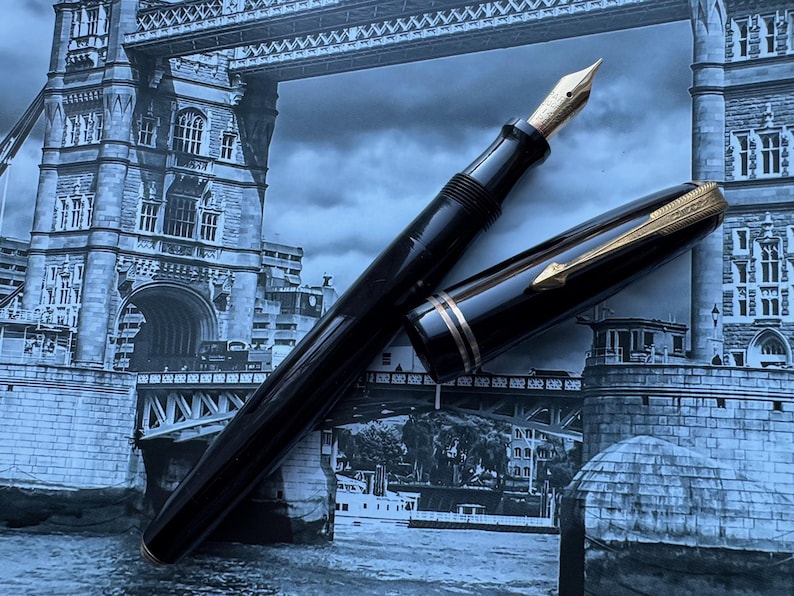 May include: A black fountain pen with a gold nib and accents, lying open on a background of the Tower Bridge in London. The pen's cap is off, revealing the nib. The pen has a sleek, classic design.