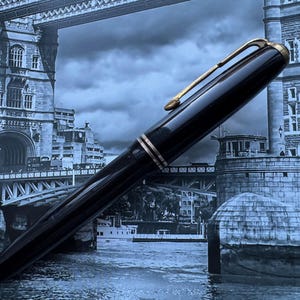 May include: A black fountain pen with gold-colored accents, including the clip and bands. The pen is set against a backdrop of a bridge and cityscape, creating a classic and elegant aesthetic.