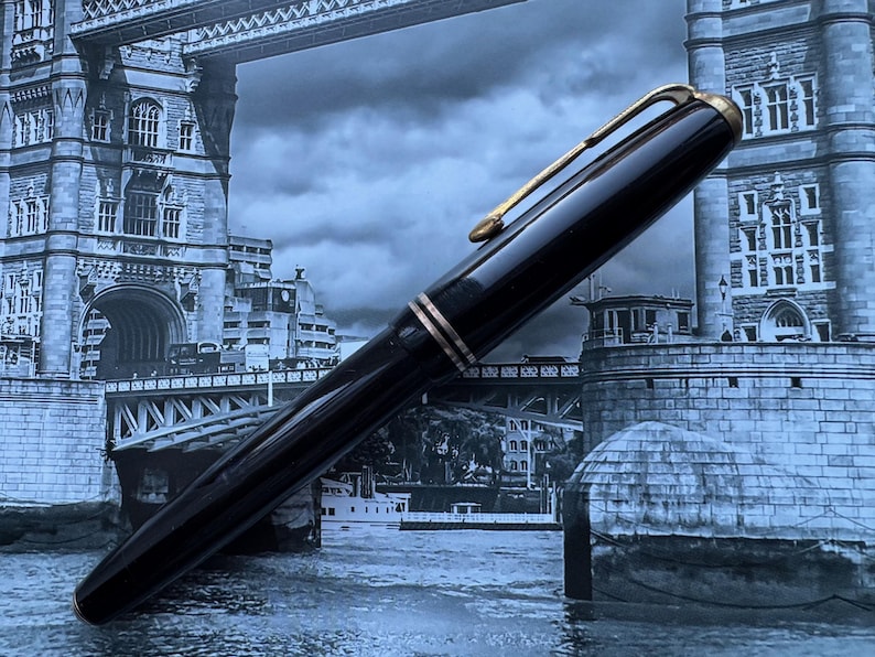 May include: A black fountain pen with gold-colored accents, including the clip and bands. The pen is set against a backdrop of a bridge and cityscape, creating a classic and elegant aesthetic.