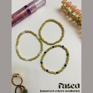 May include: Three gold beaded bracelets with varying accents, including pearls and green beads, are displayed on a white surface. A tube of lip gloss, a gold hair clip with pink flowers, and two rings are also visible. The word "Fated" is written in a stylized font.