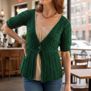 May include: A green crochet cardigan with short sleeves and a single button closure. The cardigan features a textured stitch pattern on the sleeves and upper bodice, with a ribbed pattern on the lower portion. The garment is worn over a light-colored top and blue jeans.