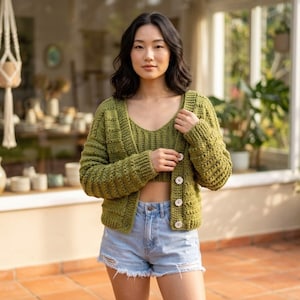 Op de afbeelding: Een groene gehaakte cardigan met een bijpassende crop top en lichtblauwe spijkerbroekshorts. De cardigan heeft een geribbelde textuur, lange mouwen en witte knopen. De outfit is een casual, handgemaakte stijl.