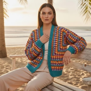 Op de afbeelding: Een kleurrijk vest met open voorkant en een geometrisch patroon. De trui heeft een turkooizen rand en is gemaakt van verschillende kleuren, waaronder oranje, paars en geel. Het vest wordt gedragen over een witte top en een beige broek. De achtergrond toont een strandscène.