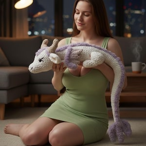 May include: A handmade crochet dragon plushie in white and lavender. The dragon has a long tail and is being held. The plushie has horns and a detailed face. The dragon is a soft toy.