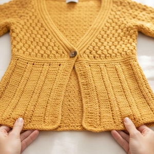 May include: Mustard yellow crochet cardigan with a V-neck and a single button fastening. The bodice features a textured stitch pattern, while the lower section has vertical ribbing. The sleeves are three-quarter length. A handmade garment.