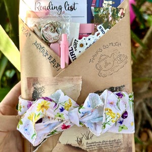 Puede incluir: Un regalo de misterio YA envuelto en papel marrón, atado con una cinta floral. El paquete contiene una lista de lectura, pegatinas, un llavero y otros pequeños artículos rosas. Las palabras "YA Mystery" y "Thanks So Much" están impresas.