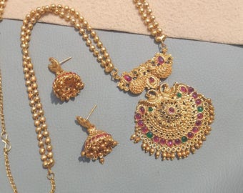 May include: A gold-toned necklace and matching jhumka earring set featuring intricate metalwork, small round beads, and inlaid pink and green stones. The necklace includes a large, ornate circular pendant. The text Majestic Fashion Hub is visible.