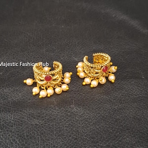 Puede incluir: Un par de pendientes tipo ear cuff dorados con un diseño de filigrana intrincado, una piedra roja central y cuentas blancas colgantes similares a perlas. El texto Majestic Fashion Hub es visible. Estos adornos circulares tienen textura.