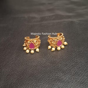 May include: A pair of gold-toned hoop earrings featuring intricate filigree metalwork, a central teardrop-shaped pink stone, and small dangling white beads along the bottom edge. The text Majestic Fashion Hub is visible above the earrings.