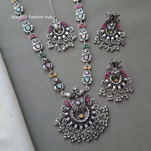 Puede incluir: Un conjunto de joyas plateadas que incluye un collar, un colgante y un par de pendientes. El collar y los pendientes tienen detalles coloridos. El collar tiene un colgante grande con detalles intrincados. El conjunto de joyas se muestra sobre una superficie gris.