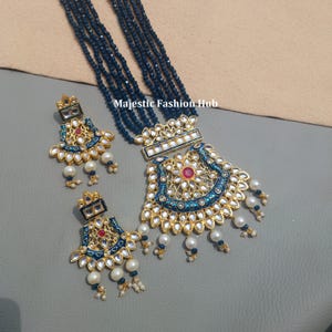 Peut inclure: Un ensemble de collier et boucles d'oreilles dorées. Le collier présente un grand pendentif avec des détails en émail bleu, une pierre rouge centrale et des perles pendantes. Les boucles d'oreilles reprennent le design du pendentif. Le collier a plusieurs rangées de perles bleu foncé.