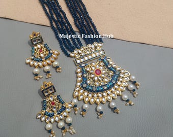 May include: An ornate gold-toned necklace and matching earrings set. The necklace features a large pendant with blue enamel detailing, a central red stone, and dangling pearls. The earrings mirror the pendant design. The necklace has multiple strands of dark blue beads.