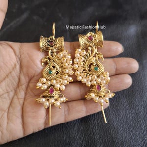 May include: A pair of gold earrings with a peacock design. The earrings have small pearls, red and green gemstones, and a decorative top. The text "Majestic Fashion Hub" is visible.
