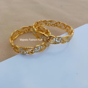 May include: Two gold-colored bangles with intricate, openwork designs. The bangles feature a repeating pattern of swirling shapes and are accented with silver-colored details. The text "Majestic Fashion Hub" is visible.