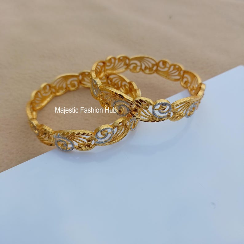 May include: Two gold-colored bangles with intricate, openwork designs. The bangles feature a repeating pattern of swirling shapes and are accented with silver-colored details. The text "Majestic Fashion Hub" is visible.