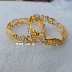May include: Two gold-colored bangles with intricate filigree designs. The bangles feature a repeating pattern of swirling details and floral accents, with silver-colored highlights. The text "Majestic Fashion Hub" is visible.