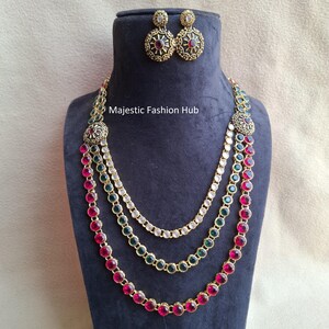 May include: A three-strand necklace and matching earrings set. The necklace features red, green, and clear gemstones, with gold-colored metal accents. The earrings are round with a similar design. The text "Majestic Fashion Hub" is visible.