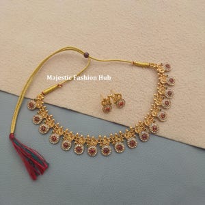 May include: Gold-coloured necklace and earring set. The necklace has circular pendants with red and clear accents. The earrings match the pendant design. The text "Majestic Fashion Hub" is visible.