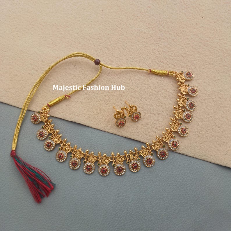 May include: Gold-coloured necklace and earring set. The necklace has circular pendants with red and clear accents. The earrings match the pendant design. The text "Majestic Fashion Hub" is visible.
