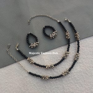 Puede incluir: Un conjunto de joyas que incluye un collar, una pulsera y un anillo. El conjunto incluye hebras de cuentas negras con detalles plateados. El anillo y la pulsera están conectados por una cadena plateada. El texto "Majestic Fashion Hub" es visible.