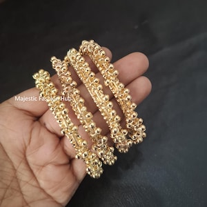May include: A set of three gold-toned bangles featuring a textured surface composed of small, spherical beads arranged in a repeating pattern. The metal surface has a reflective, polished finish. The text Majestic Fashion Hub is visible on the left.