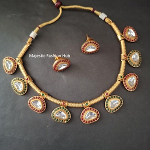 May include: A gold-toned necklace and matching stud earrings set featuring triangular clear stones bordered by small red and green gems. The necklace has a textured gold cord and the text Majestic Fashion Hub is visible.