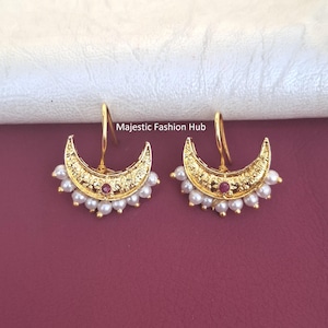 Op de afbeelding: Een paar goudkleurige oorbellen in de vorm van een halve maan, met een ingewikkelde reliëftextuur, een kleine centrale rode steen en een rij witte parels langs de onderrand. De tekst Majestic Fashion Hub is in het midden zichtbaar.