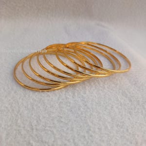 May include: A collection of gold-colored bangles, arranged in a slightly overlapping pattern. The bangles are thin and circular, reflecting light. The background is a soft, white fabric. The text "Majestic Fashion Hub" is visible in the upper left corner.