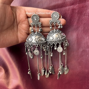 May include: Silver-toned, ornate earrings with a bell-shaped design. Each earring features a floral top, dangling chains, small bells, leaf-shaped charms, and coin-like pendants. The earrings are displayed against a burgundy background.