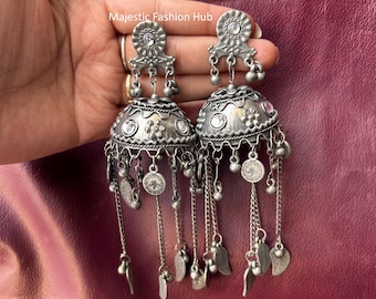 May include: Silver-toned, ornate earrings with a bell-shaped design. Each earring features a floral top, dangling chains, small bells, leaf-shaped charms, and coin-like pendants. The earrings are displayed against a burgundy background.