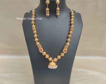 May include: Gold-toned necklace and earring set. The necklace features a beaded design with a pendant and red and green accents. The earrings have a similar beaded design. The jewelry is displayed on a black mannequin.