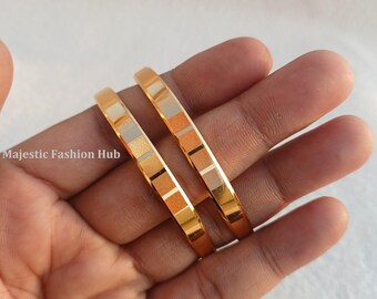 Könnte beinhalten: Zwei goldfarbene Armreifen mit einem quadratischen Muster. Die Armreifen werden in einer Hand gehalten. Das quadratische Muster wechselt zwischen Gold-, Silber- und Kupfertönen. Der Text "Majestic Fashion Hub" ist im Bild sichtbar.