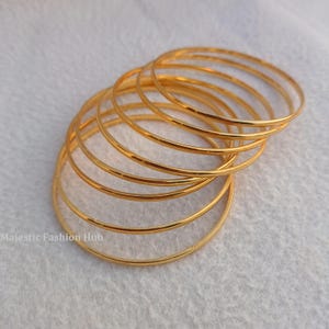 May include: A collection of gold-colored bangles, arranged in a semi-circular pattern. The bangles are thin and have a smooth, reflective surface. The image is set against a soft, light-colored background. The text "Majestic Fashion Hub" is visible in the lower left corner.