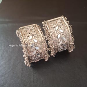 Puede incluir: Un par de brazaletes de metal plateado con un intrincado patrón de hojas incrustado con pequeños espejos reflectantes. Los bordes están adornados con delicados bucles de alambre y cuentas metálicas colgantes. El texto Majestic Fashion Hub es visible.