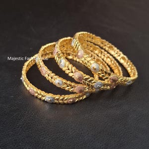 Könnte beinhalten: Ein Set aus drei goldfarbenen Armreifen mit einem sich wiederholenden Blattmuster, das mit kreisförmigen Akzenten in Silber und Roségold durchsetzt ist. Der Text Majestic Fashion Hub ist links auf den strukturierten Armreifen zu sehen.