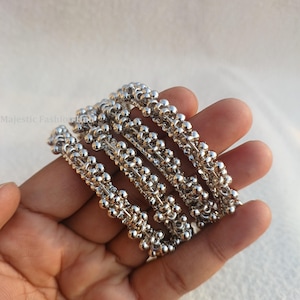 May include: Four silver bangles with a beaded design. Each bangle is made up of small, round beads linked together. The bangles are displayed on a hand, highlighting their intricate details and reflective surface.